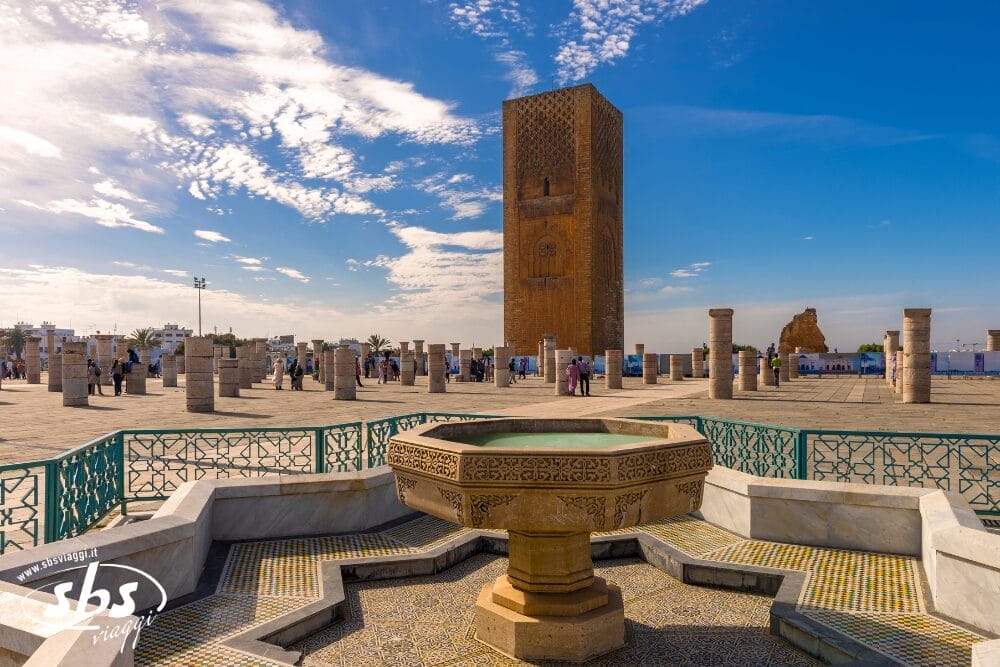 Una fontana in pietra con piastrelle a mosaico si trova in primo piano, mentre l'alta torre di Hassan, incompiuta, e le colonne sparse si ergono nella piazza soleggiata - una scena iconica spesso catturata attraverso Bozza automatica sotto un cielo parzialmente nuvoloso.