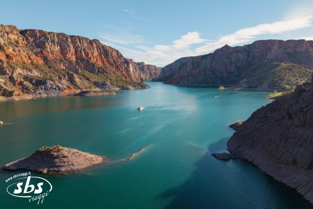 Un ampio fiume turchese scorre tra scogliere rocciose e rossastre e colline verdi sotto un cielo parzialmente nuvoloso. Una piccola barca è visibile sull'acqua. Il logo "sbs viajes" e la filigrana Bozza automatica appaiono nell'angolo in basso a sinistra.