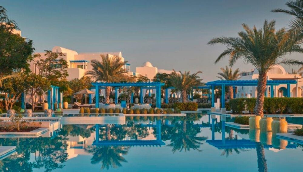 Un lussuoso resort sulla spiaggia con edifici bianchi, pergolati blu, palme e vegetazione lussureggiante circonda una grande e tranquilla piscina che riflette il tramonto: l'ideale per abbracciare lo stile Seaclub dell'Hilton Salwa Beach Resort.
