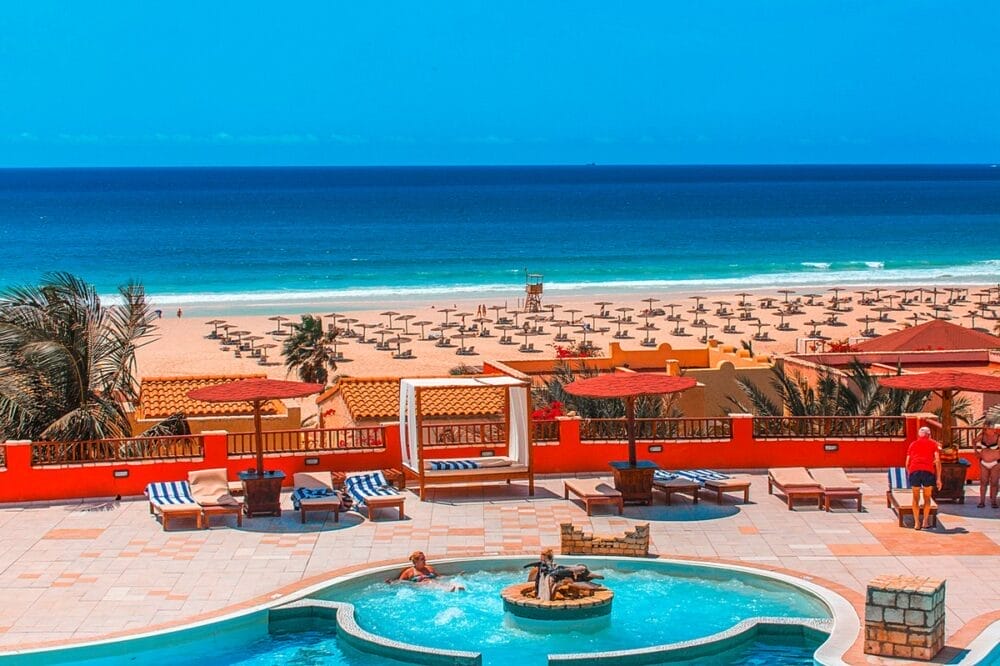 La vivace area della piscina dell'Occidental Boa Vista, con sedie a sdraio e ombrelloni, si affaccia sulle spiagge sabbiose di Boa Vista Beach, fiancheggiate da lettini, palme e l'oceano blu brillante sotto un cielo limpido.