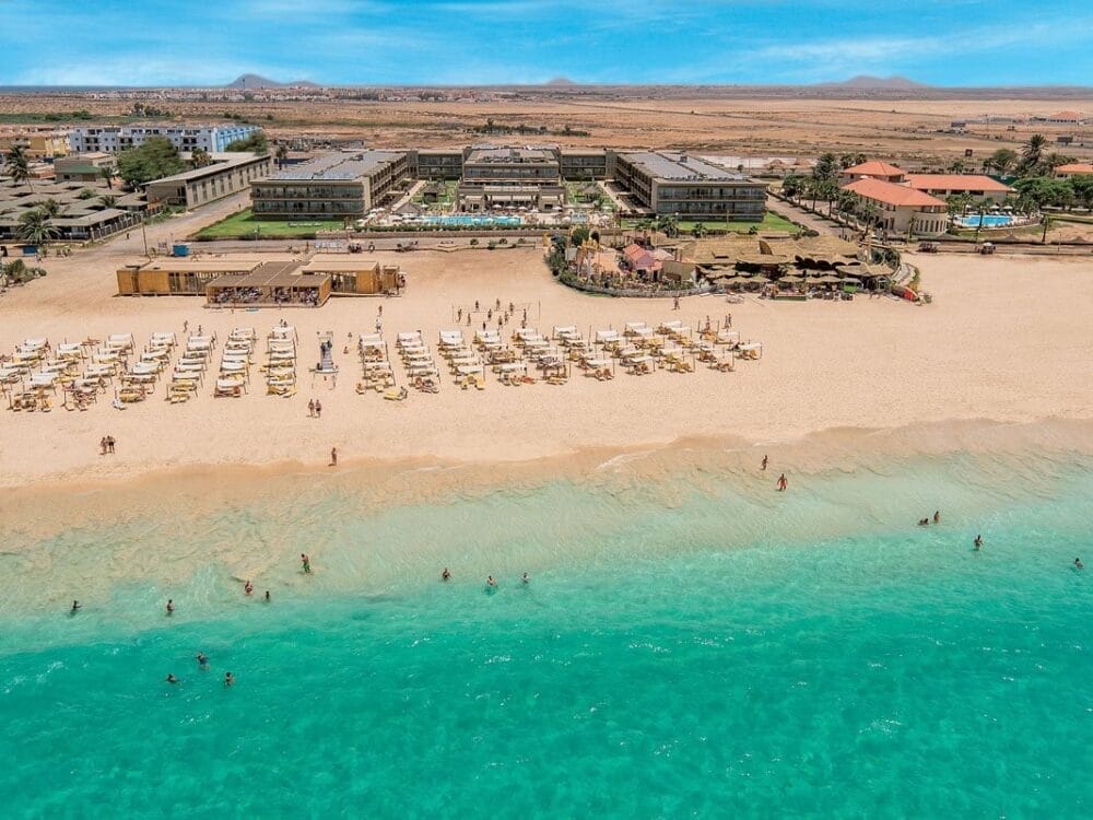 Vista aerea del resort Veraclub Oasis Salinas Sea sulla spiaggia, con file di lettini e ombrelloni sulla sabbia dorata, persone che nuotano nell'acqua turchese ed edifici con piscine e giardini dietro la spiaggia.