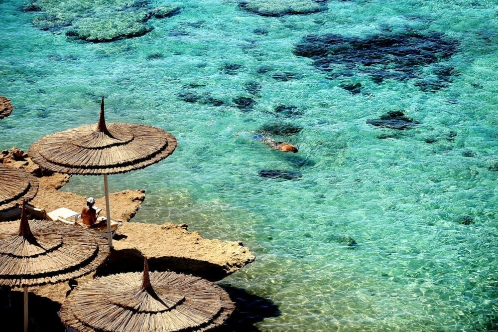Una persona si rilassa su una sedia a sdraio sotto un ombrellone di paglia al Reef Oasis Beach Resort, mentre un'altra nuota in acque turchesi e limpide vicino ad affioramenti rocciosi. La scena illuminata dal sole è caratterizzata dalle ombre degli ombrelloni che danzano sulle rocce.