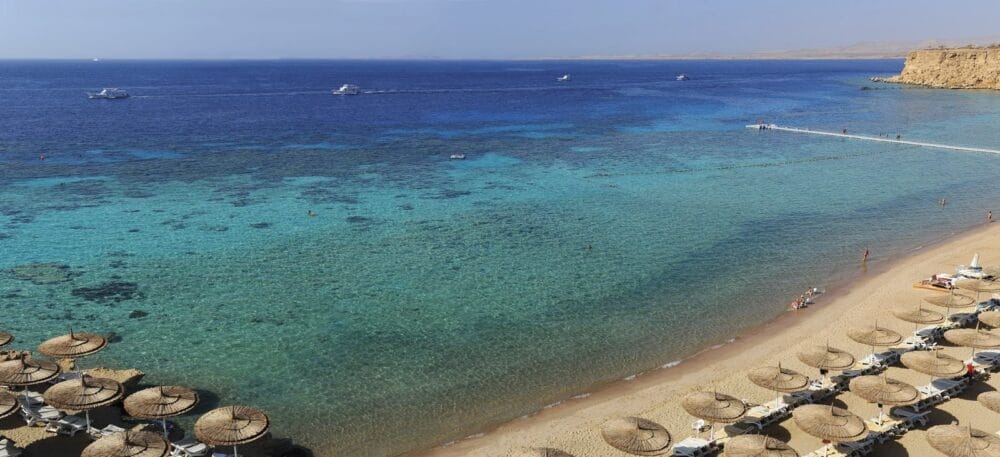 Una spiaggia sabbiosa con file di ombrelloni di paglia, un'acqua limpida e turchese e alcune barche sparse sul mare calmo e blu sotto un cielo limpido, a pochi passi dal comfort rilassante del Reef Oasis Senses.