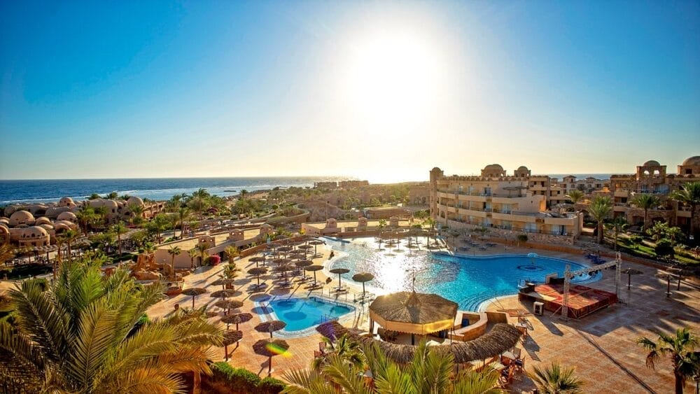 Un resort illuminato dal sole con una grande piscina all'aperto, palme, sedie a sdraio e ombrelloni si affaccia sul mare del Veraclub Utopia Beach di Marsa Alam. Edifici con tetti a cupola circondano la piscina, sotto un sole splendente e un cielo azzurro e limpido.