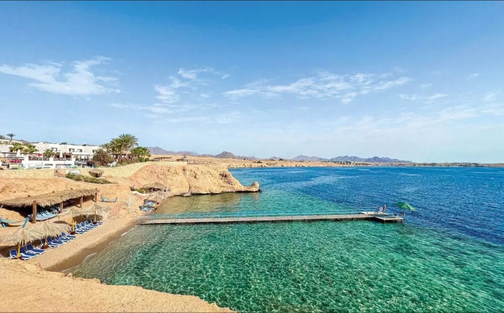 La vista panoramica della costa del Veraclub Sharm mostra un'acqua limpida e turchese, un molo di legno che si protende nel mare, scogliere sabbiose e lettini sotto ombrelloni di paglia; edifici del resort e palme costeggiano la riva di Sharm el Sheikh sotto un cielo azzurro e luminoso.