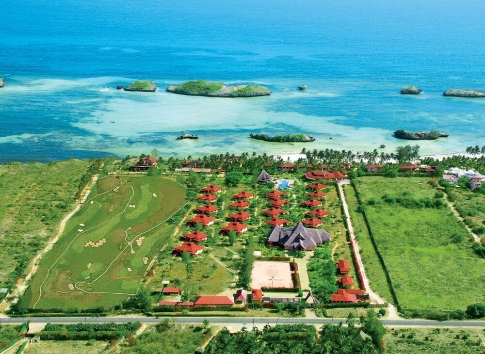 Vista aerea del resort costiero Veraclub Crystal Bay, con ville dai tetti rossi, prati verdi, giardini, piscina e campo da golf, delimitato da una vegetazione lussureggiante e affacciato su un oceano turchese con piccole isole al largo.