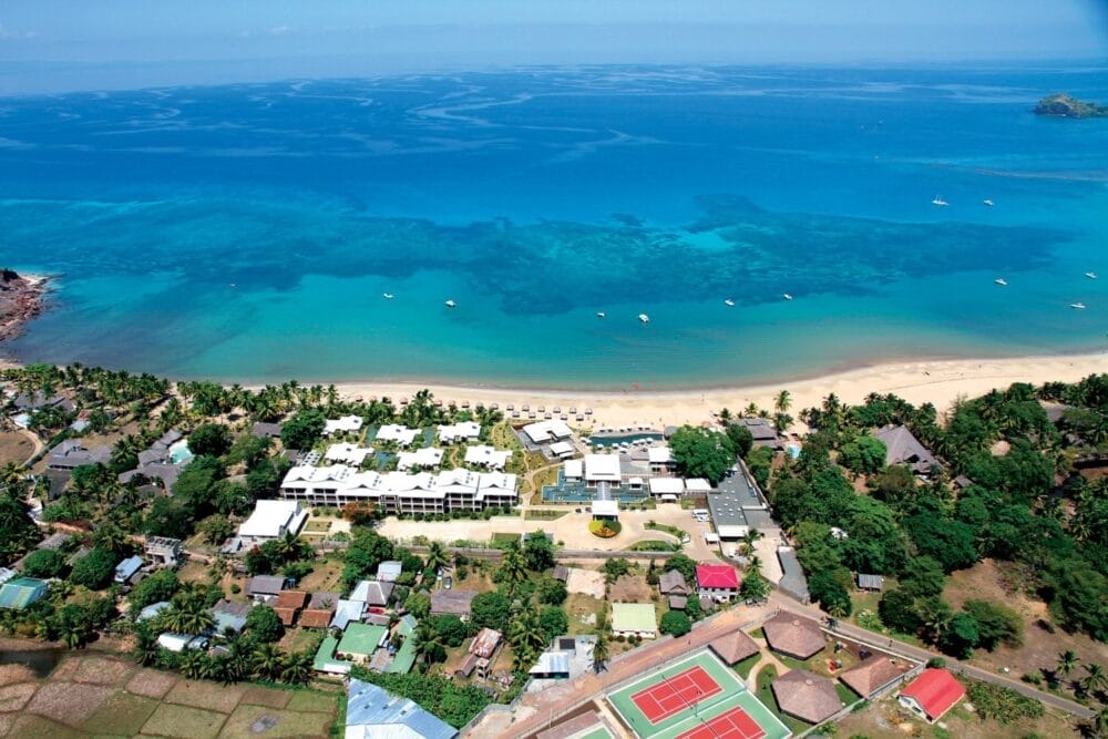 Veduta aerea del resort Veraclub Palm Beach con sabbia bianca, acqua cristallina, edifici sulla spiaggia, palme, campi da tennis e barche ancorate al largo. Una vegetazione lussureggiante e le strutture del villaggio circondano il resort.
