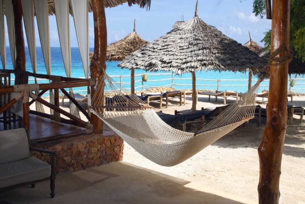 Un'amaca intrecciata è appesa tra pali di legno vicino a un patio ombreggiato sulle rive sabbiose del Veraclub Sunset Beach, con ombrelloni di paglia, sedie a sdraio, mare turchese e cielo azzurro sullo sfondo.
