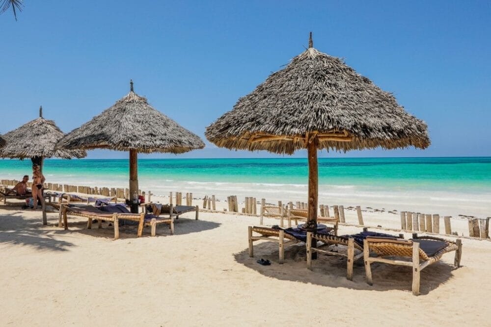 Ombrelloni di paglia e sedie a sdraio in legno su una spiaggia sabbiosa del Veraclub Zanzibar, di fronte all'acqua turchese dell'oceano sotto un cielo azzurro e limpido. Alcune persone si rilassano all'ombra vicino alla riva, immergendosi nella tranquilla atmosfera del villaggio di Zanzibar.