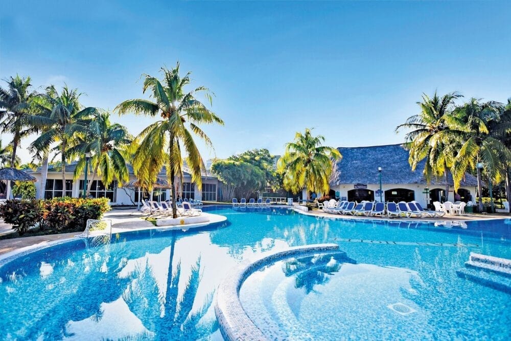 La piscina di un hotel tropicale a Cuba è circondata da palme, lettini e tetti di paglia sotto un cielo azzurro e limpido. La grande piscina del Veraclub Starfish Varadero è caratterizzata da bordi curvi e aree di seduta integrate per un perfetto relax.