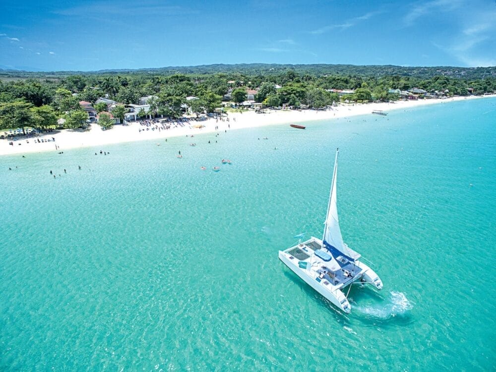 Un catamarano bianco naviga su acque limpide e turchesi vicino a una spiaggia sabbiosa fiancheggiata da alberi ed edifici, dove gli ospiti del Veraclub Negril si divertono a nuotare e a rilassarsi sulla riva sotto un cielo azzurro e luminoso.
