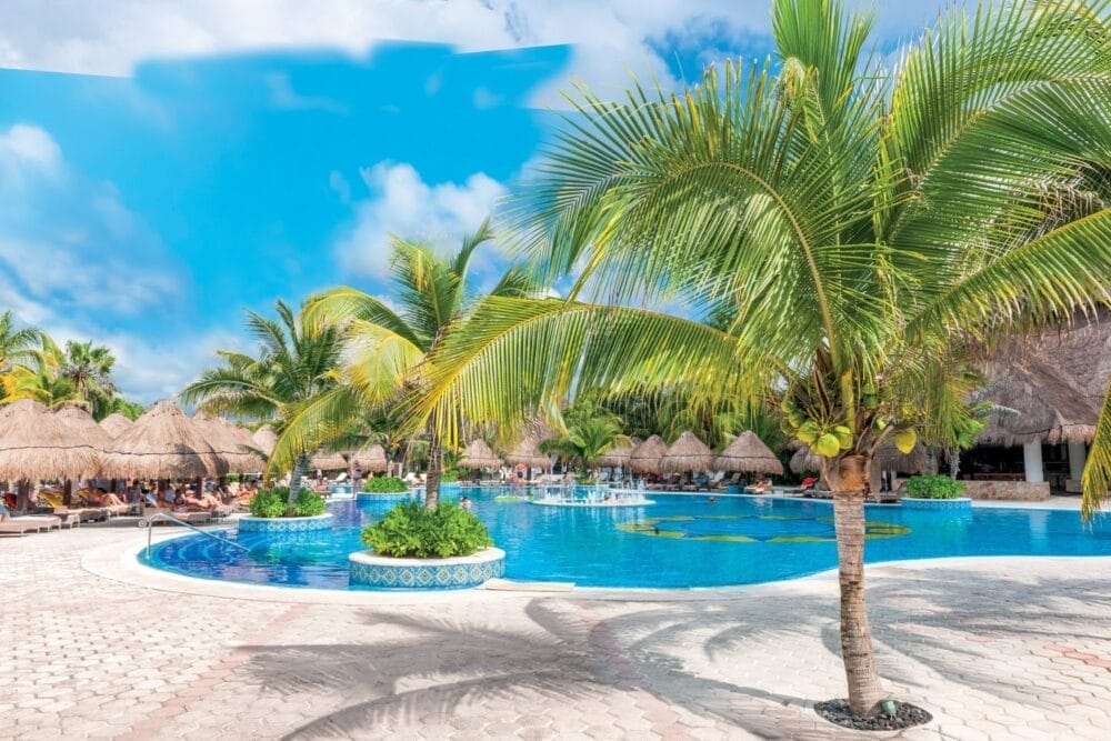 La piscina del resort tropicale Veraclub Royal Tulum è circondata da palme, sedie a sdraio e capanne con tetto di paglia sotto un cielo blu brillante. Questo resort di Tulum ha un aspetto solare, invitante e perfetto per rilassarsi in un hotel all-inclusive.