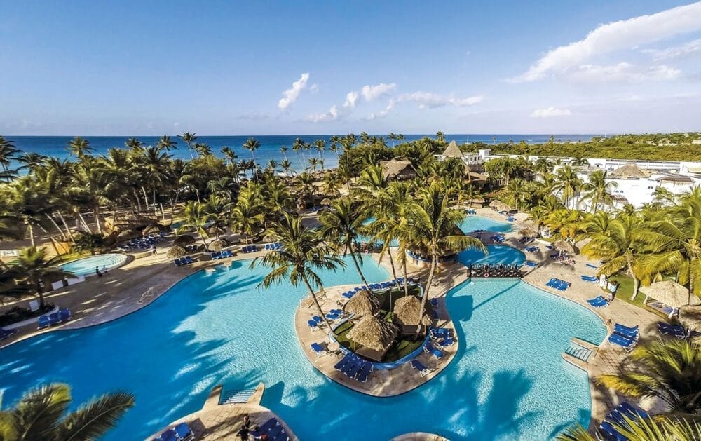 Vista aerea del Sunscape Dominicus, un resort tropicale con grandi piscine, palme, lettini e capanne con tetto di paglia vicino alla spiaggia e all'oceano blu, sotto un cielo soleggiato. Ideale per una rilassante fuga Veraclub.