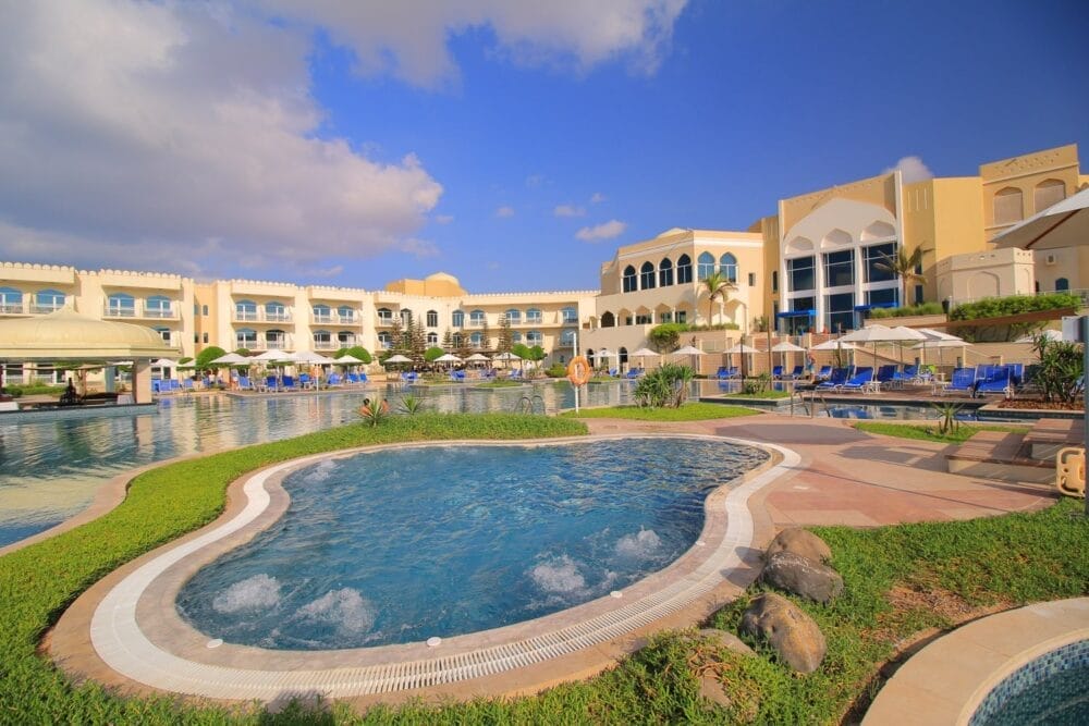 Una scena di resort al Veraclub Salalah con una grande piscina, una vasca idromassaggio rotonda in primo piano, sedie a sdraio, ombrelloni, palme e un edificio giallo e bianco in stile mediterraneo sotto un cielo parzialmente nuvoloso.
