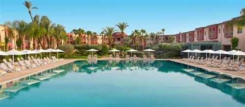 Una grande piscina all'aperto è circondata da sedie a sdraio e ombrelloni, con palme e il resort Iberostar Palmeraie Marrakech rosa a due piani sullo sfondo, sotto un cielo azzurro e limpido.