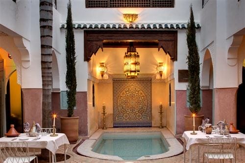 Un elegante cortile marocchino della Angsana Riads Collection è caratterizzato da una piccola piscina centrale, da un intricato mosaico di piastrelle, da lanterne decorate, da alti alberi in vaso e da tavoli da pranzo a lume di candela. L'illuminazione calda crea un'atmosfera accogliente da Angsana Hotels.