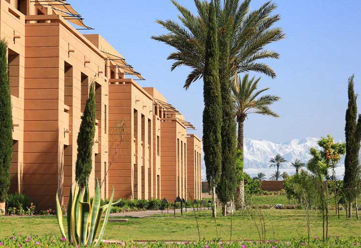 Un edificio moderno con pareti color marrone e finestre alte e strette, che ricorda l'Aqua Mirage Marrakech, è circondato da palme e cipressi, erba verde in primo piano e montagne innevate sotto un cielo azzurro.