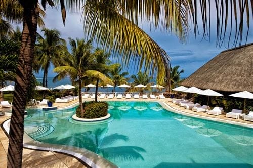 Una lussuosa piscina di un resort di Mauritius circondata da palme, lettini con ombrelloni bianchi e un edificio con tetto di paglia, che si affaccia su un oceano calmo sotto un cielo parzialmente nuvoloso: una fuga idilliaca al Maritim Resort.