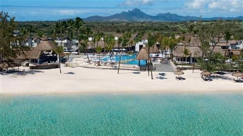 Una vista della spiaggia tropicale dell'Ambre Sun Resort con sabbia bianca, acqua turchese e trasparente, edifici con tetti di paglia, palme, piscine e lontane montagne verdi sotto un cielo azzurro.