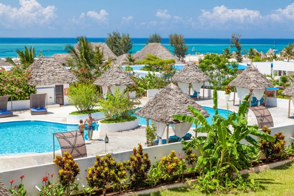 Il Bravo Kendwa Beach Resort è un paradiso tropicale con cabine dal tetto di paglia, piscine, palme e giardini lussureggianti che si affacciano sull'oceano turchese sotto un cielo azzurro e soleggiato. Gli ospiti si rilassano e nuotano in piscina, godendosi ogni momento.