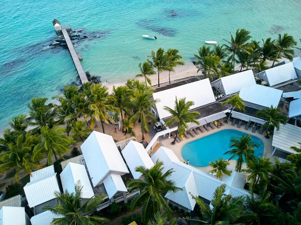 Veduta aerea di Tropical Attitude, un resort balneare per soli adulti a Mauritius, con edifici dai tetti bianchi, palme, una piscina curva e un molo di legno che si protende nelle acque turchesi. Una piccola barca galleggia vicino alla riva.