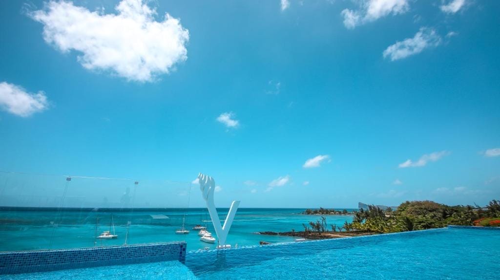 La piscina a sfioro dell'Ocean V Hotel si affaccia su un oceano turchese punteggiato di barche ancorate, su un cielo limpido e su una costa tropicale dalla vegetazione lussureggiante. Una scultura bianca si erge elegantemente sul bordo della piscina.