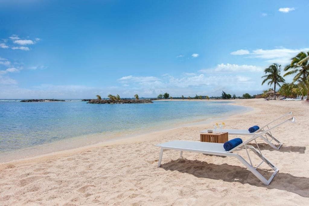 Due sedie a sdraio bianche con asciugamani blu e bevande si trovano sulla spiaggia sabbiosa del Veranda Pointe Aux Biches, un tranquillo resort balneare a Mauritius. Le palme costeggiano la riva sotto un cielo soleggiato con nuvole sparse.