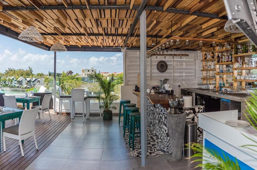 Il bar e ristorante sul tetto, Azur Paradise, presenta un soffitto in legno, un arredamento moderno, un bancone con piastrelle a fantasia, luci pendenti, sedie bianche e verde acqua, piante in vaso e una splendida vista sugli edifici e sul cielo sullo sfondo.