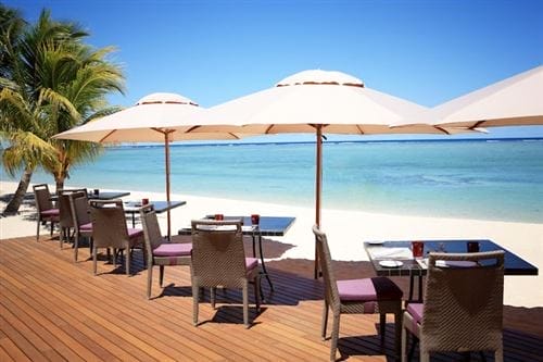 Zona pranzo all'aperto con tavoli e sedie sotto grandi ombrelloni bianchi su un ponte di legno presso la spiaggia sabbiosa del Lux Le Morne, con vista sulle acque azzurre dell'oceano e un cielo luminoso e soleggiato. Una palma è visibile sulla sinistra.