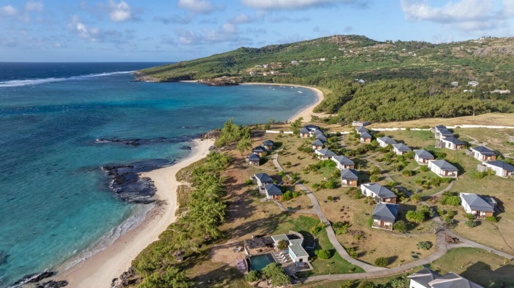 Vista aerea di un paesaggio costiero panoramico con onde turchesi, spiagge di sabbia bianca, verde sparso e cottage o ville Constance Tekoma Rodrigues vicino alla costa, delimitato da dolci colline sotto un cielo parzialmente nuvoloso.