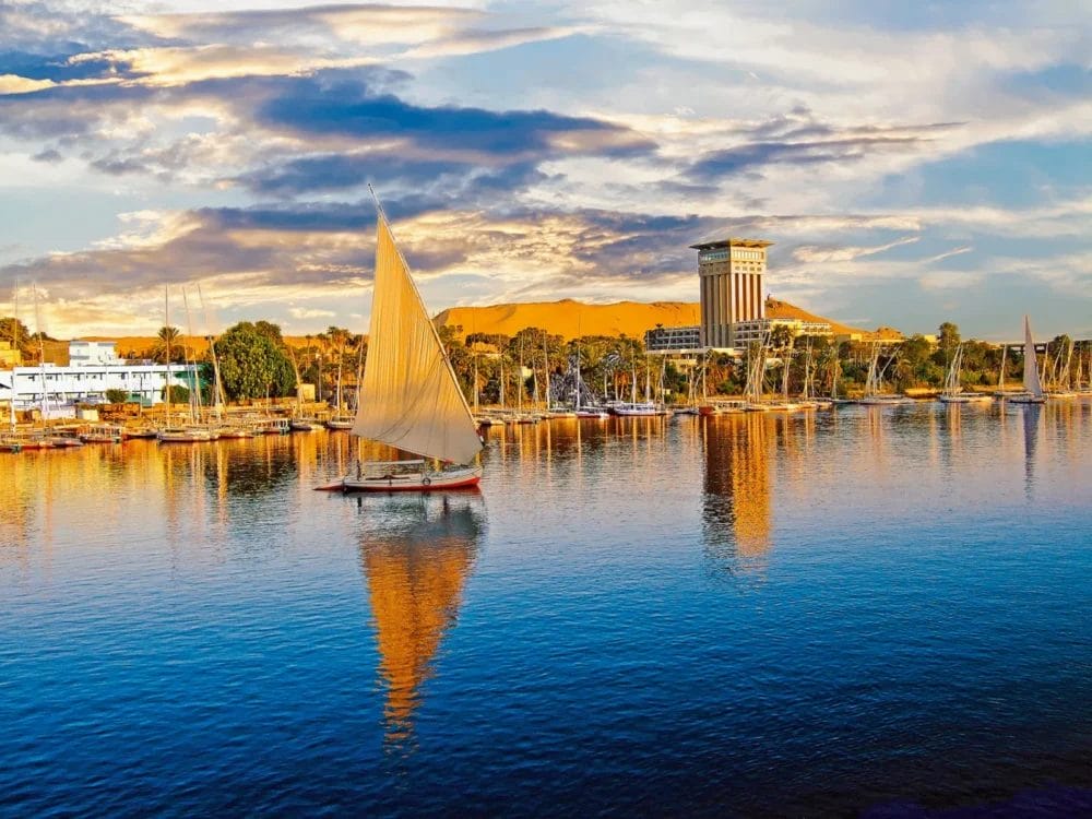 Una barca a vela tradizionale scivola su un fiume calmo al tramonto vicino a Il Cairo, con riflessi sull'acqua, alberi che costeggiano la riva, un alto edificio moderno e colline dorate sotto un cielo drammatico e pieno di nuvole: l'ideale per una Crociera Nefertari.