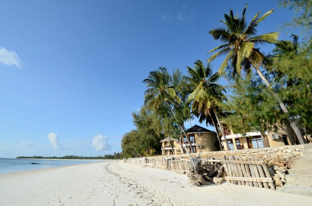Una spiaggia sabbiosa con cielo azzurro e limpido, palme e cottage sulla spiaggia circondati dal verde a Kae Beach Zanzibar. Il litorale si estende in lontananza con acque calme e poco profonde e nessuna persona visibile.