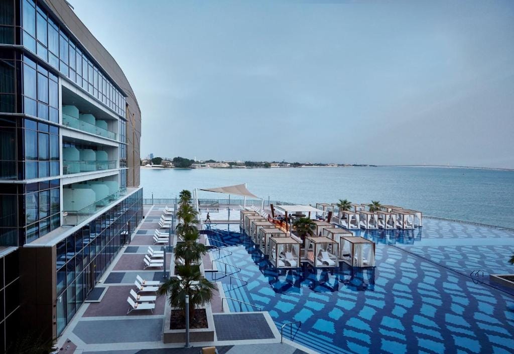 Il Royal M Hotel di Abu Dhabi dispone di moderni balconi in vetro che si affacciano su una piscina all'aperto con cabine e sedie a sdraio, il tutto in riva a un tranquillo specchio d'acqua sotto un cielo limpido.