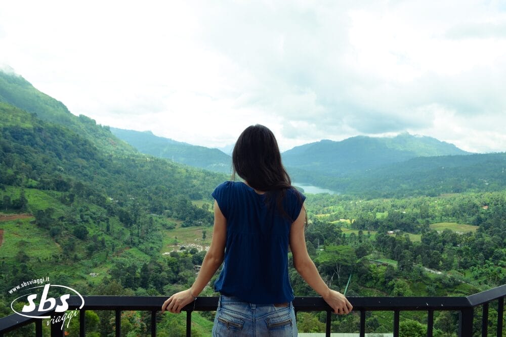 Una donna dai lunghi capelli scuri è in piedi su una ringhiera, affacciata su una valle verde e lussureggiante e sulle montagne sotto un cielo nuvoloso. Il paesaggio vibrante sembra infinito. Il logo "sbs" e "Bozza automatica" appaiono nell'angolo in basso a sinistra.