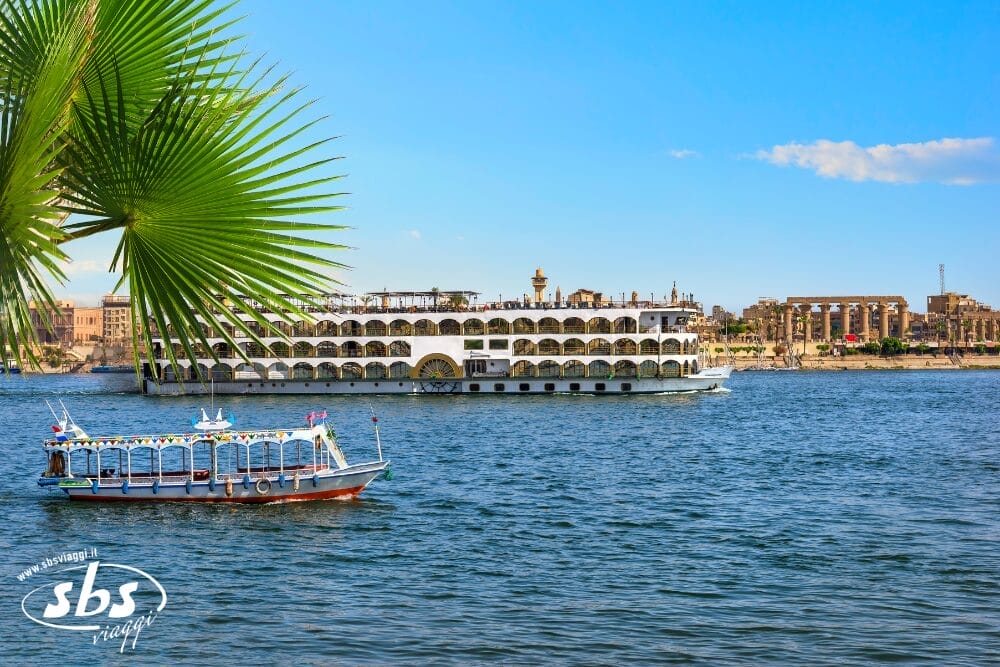 Una grande nave da crociera fluviale e una piccola imbarcazione decorata navigano sul fiume Nilo, con foglie di palma in primo piano e antiche rovine egizie visibili sulla sponda opposta sotto un cielo azzurro e limpido: una serena Bozza automatica di una bellezza senza tempo.
