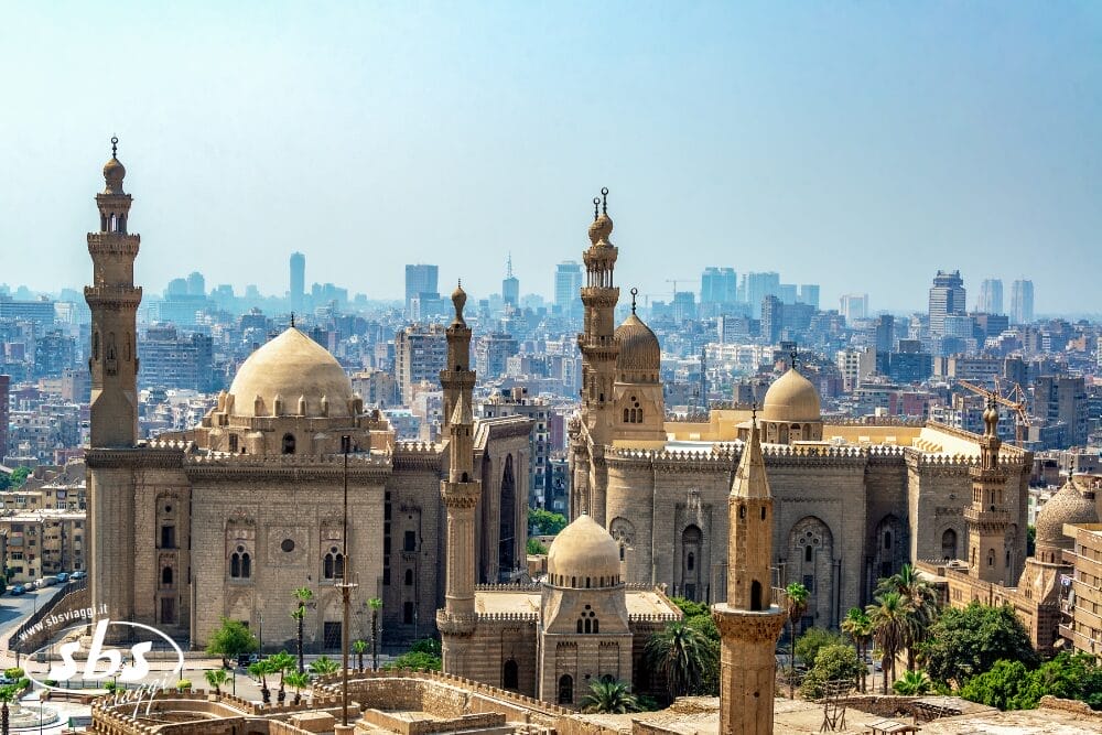Una vista panoramica di moschee storiche con grandi cupole e minareti al Cairo, in Egitto, si staglia contro lo skyline di una città moderna e un cielo azzurro e limpido: una scena perfetta per una Bozza automatica.