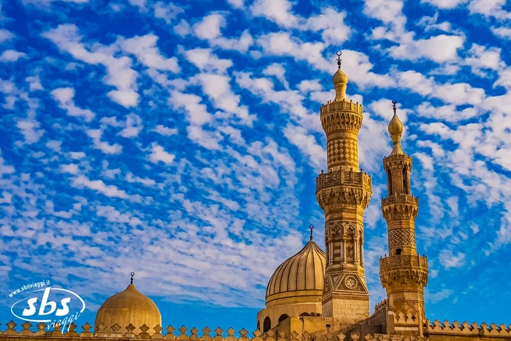 Due minareti ornati e la cupola di una moschea si stagliano su un cielo blu vibrante pieno di nuvole bianche sparse e soffici. In primo piano, una recinzione decorativa si erge mentre la luce del sole mette in risalto gli intricati dettagli della moschea, come una bozza automatica in architettura.