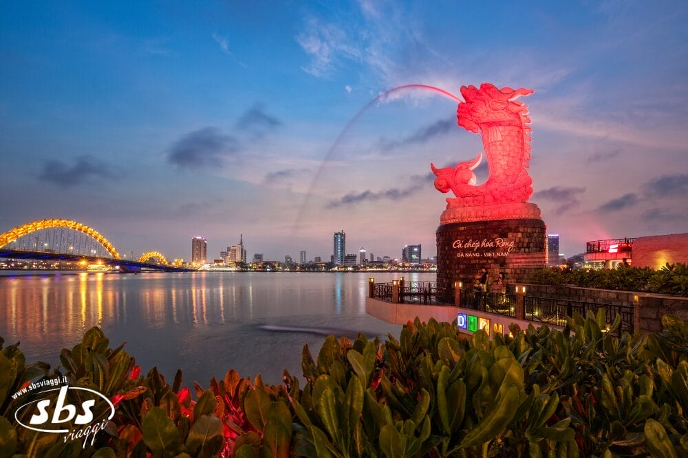 Una grande statua di drago spruzza acqua nel fiume al tramonto a Da Nang, in Vietnam. Sullo sfondo appaiono gli edifici della città e un ponte illuminato di giallo, con le piante in primo piano: una scena di vita vibrante e di bozza automatica.