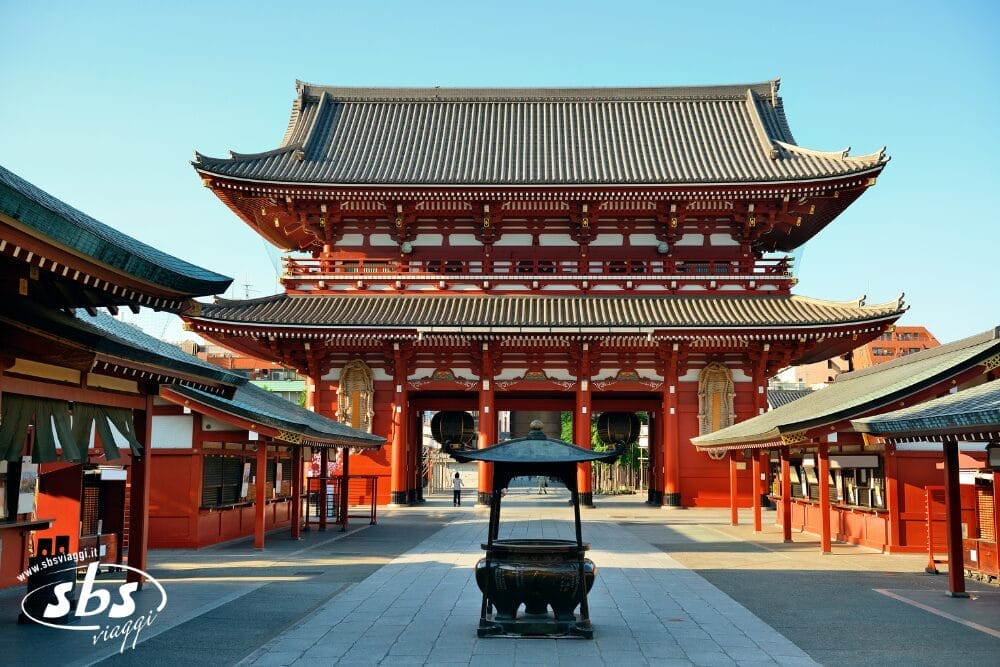Grande tempio tradizionale giapponese con struttura in legno rosso, tetto decorato e bruciatore di incenso davanti. Il cielo azzurro e il cortile vuoto creano una scena tranquilla. Il logo "Sbs viaggi" e "Bozza automatica" appaiono nell'angolo in basso a sinistra.