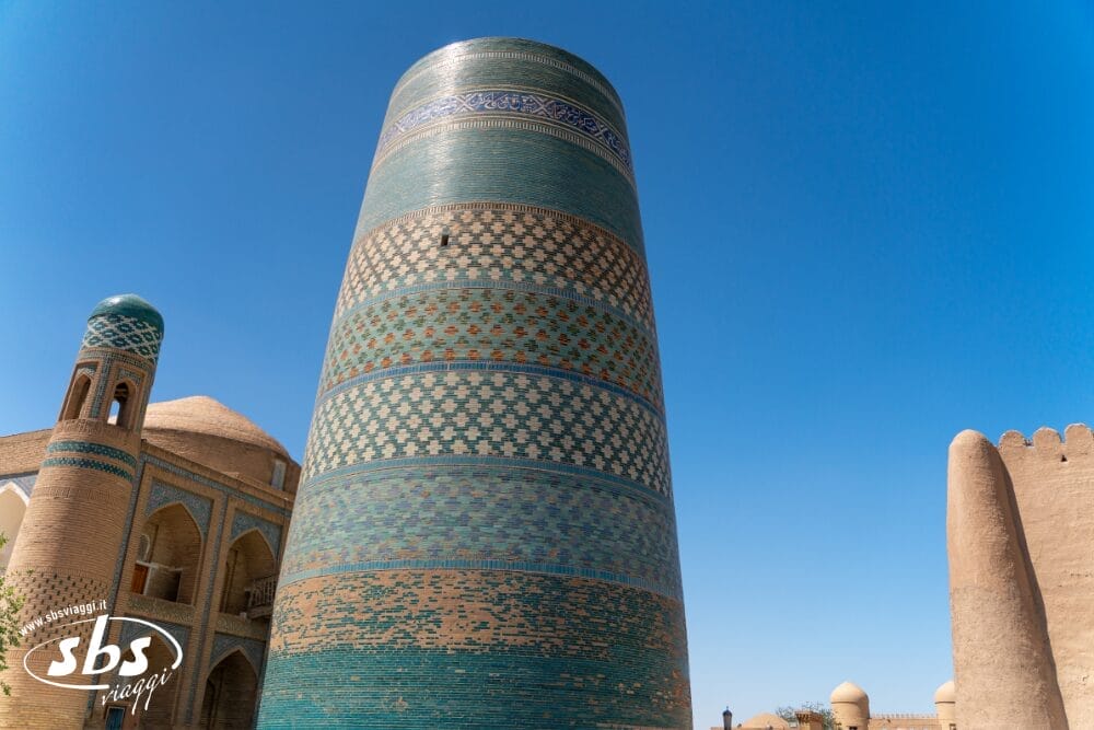 Un grande minareto cilindrico dalle piastrelle turchesi e blu, noto come Bozza automatica, si erge sotto un cielo azzurro e limpido, circondato da edifici storici color sabbia a Khiva, in Uzbekistan.