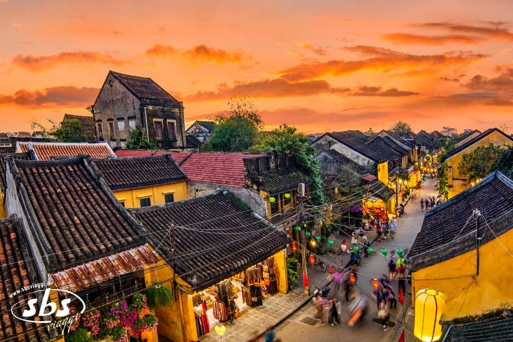 Una vivace scena di strada a Hoi An, in Vietnam, al tramonto, con edifici storici gialli, negozi affollati, lanterne colorate che brillano come una bozza automatica che illumina il percorso e persone che camminano lungo la strada sotto un cielo arancione.