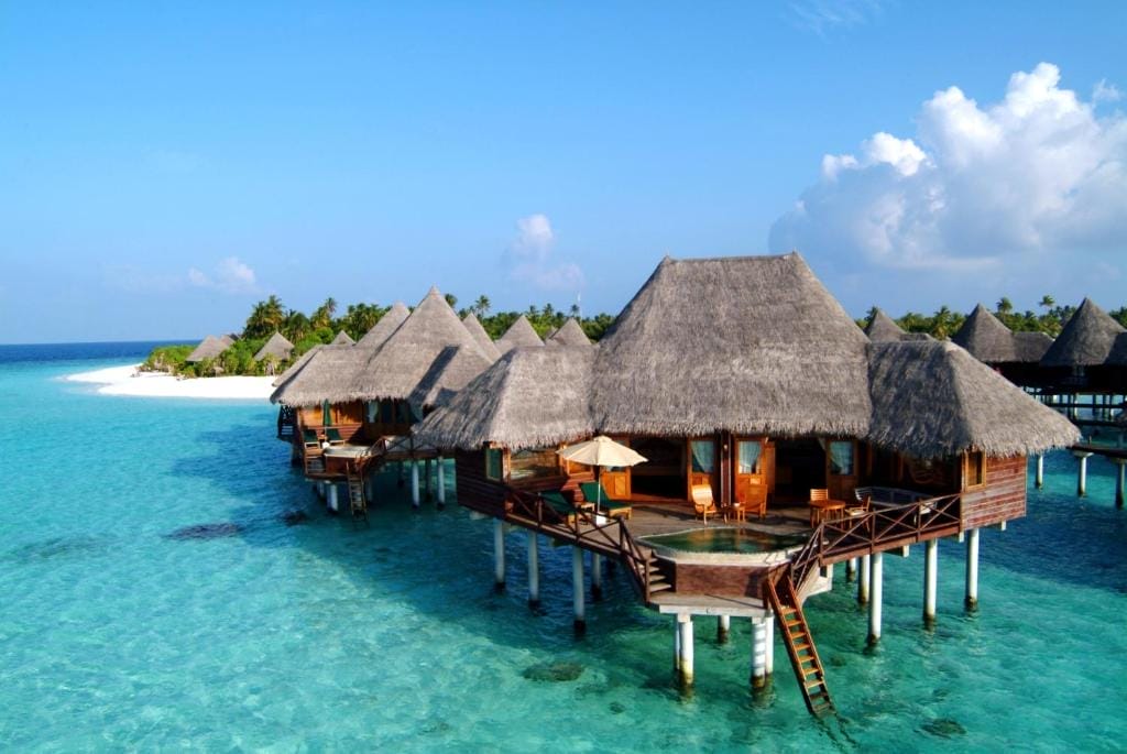 I bungalow overwater con tetti di paglia si ergono sull'acqua limpida e turchese del resort di lusso Coco Palm Dhuni Kolhu, circondato da spiagge di sabbia bianca e da una vegetazione lussureggiante sotto un cielo blu brillante con nuvole sparse.
