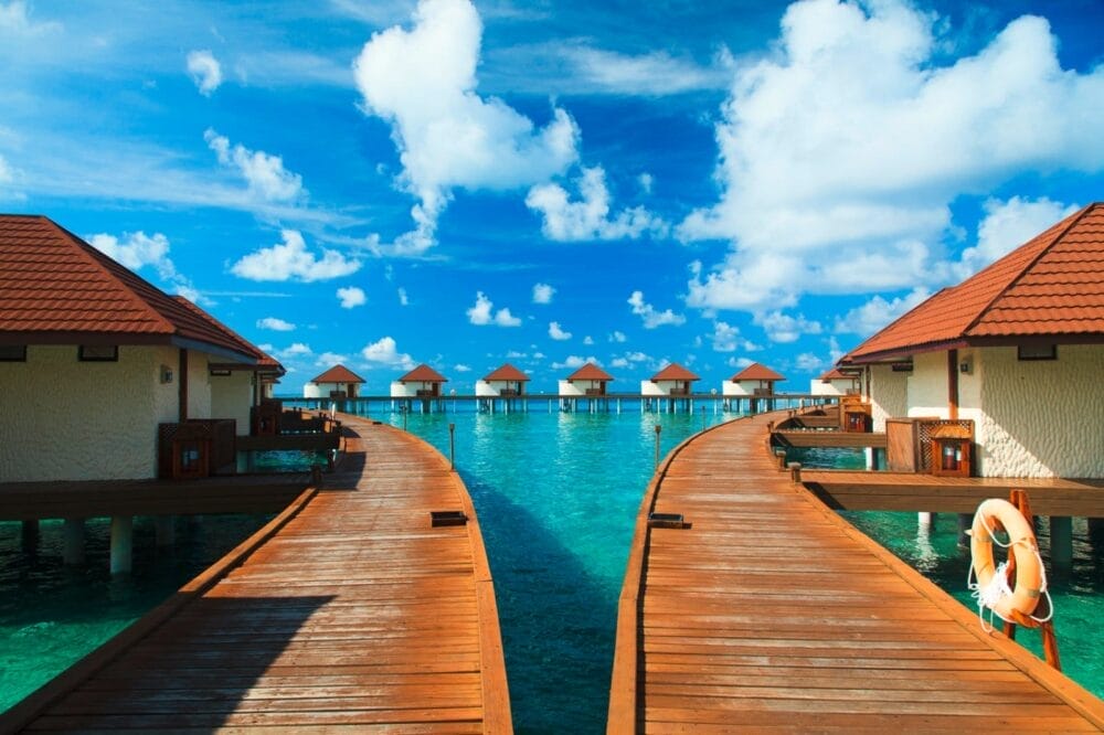 I bungalow overwater con tetti rossi e passerelle in legno del Bravo Alimatha' si estendono su acque limpide e turchesi sotto un cielo blu brillante con nuvole soffici.