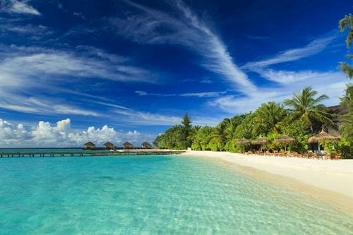 La spiaggia tropicale di Bravo Maayafushi è caratterizzata da acque turchesi e trasparenti, sabbia bianca, palme, capanne di paglia e un molo di legno, sotto un cielo blu intenso con nuvole sottili.