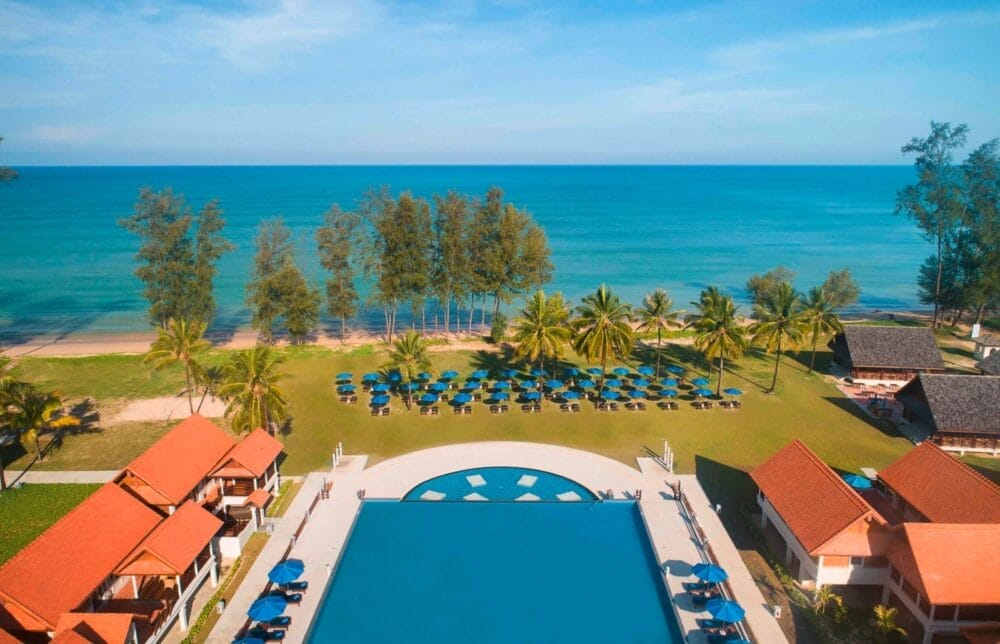 Vista aerea del resort Bravo Premium Khao Lak sulla spiaggia con una grande piscina, file di sedie a sdraio e ombrelloni blu, palme, area erbosa e l'oceano sullo sfondo sotto un cielo azzurro e limpido.