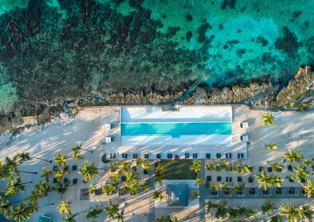 Vista aerea della piscina rettangolare del Bravo Viva Dominicus Beach vicino all'oceano turchese, circondata da sabbia bianca, palme e sedie a sdraio su una spiaggia tropicale.