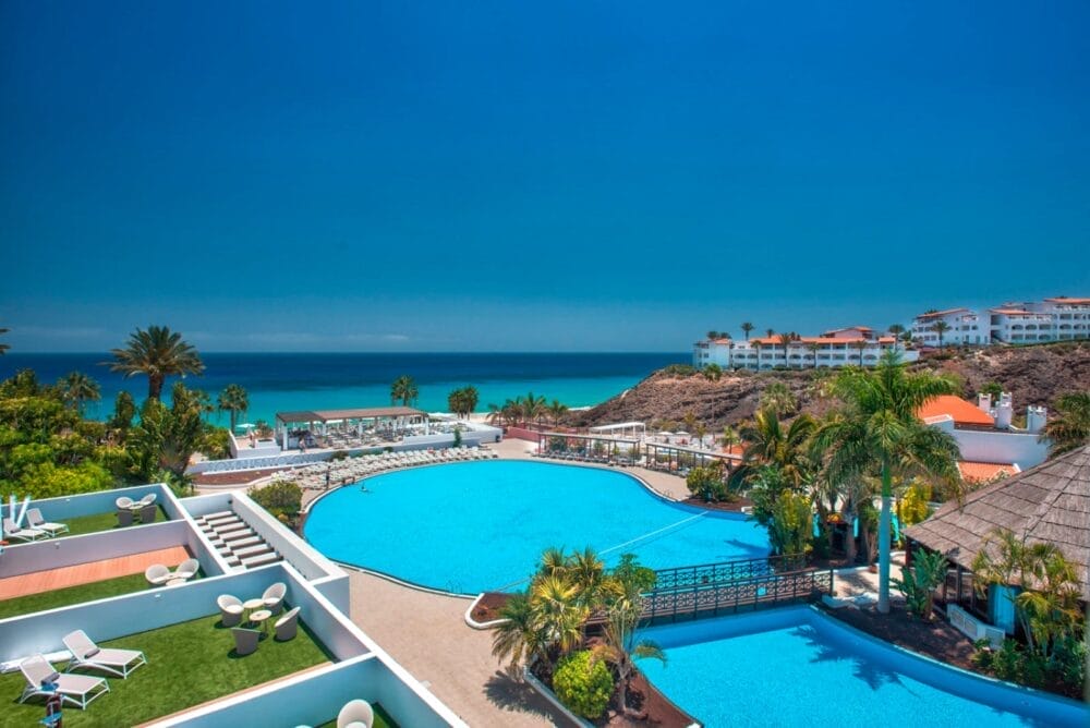 La grande piscina circolare del Bravo Fuerteventura è circondata da palme e sedie a sdraio e si affaccia su un oceano turchese sotto un cielo azzurro e limpido, con edifici e giardini bianchi sullo sfondo.