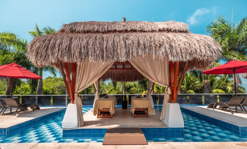 Al Bravo Decameron Baru una cabina con tetto di paglia, tende e due lettini per massaggi si trova sopra la piscina piastrellata di blu, circondata da palme, sedie a sdraio e ombrelloni rossi sotto un cielo soleggiato.