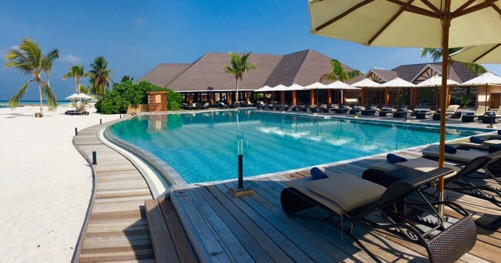 La piscina del Brennia Kottefaru, un lussuoso resort delle Maldive, è dotata di sedie a sdraio e ombrelloni su un ponte di legno che si affaccia su una spiaggia di sabbia bianca e palme sotto un cielo azzurro e limpido.