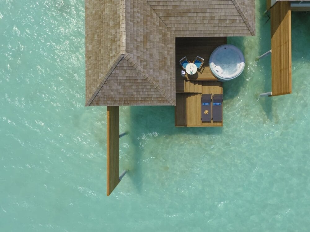 Vista aerea di un bungalow overwater in un resort delle Maldive, con una terrazza in legno con due sedie a sdraio, un tavolino per due persone e una vasca idromassaggio rotonda, il tutto circondato da acque turchesi e trasparenti.